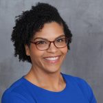 Melissa Williams wearing glasses and a blue shirt on a gray background