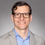 Portrait of John Jay wearing glasses, light blue shirt and gray woven jacket on a gray background.